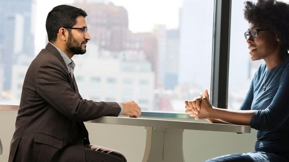 Riham Moin sitting across a table from a client by a large city window, in conversation